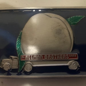 May include: A silver belt buckle with a blue background featuring a truck hauling a giant peach with the words "The Allman Brothers Band" on the side of the truck.