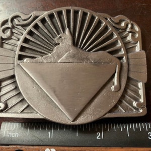 Könnte beinhalten: Eine silberfarbene Gürtelschnalle mit einem stilisierten Katzendesign in einem Dreieck. Die Katze blickt nach links und das Dreieck ist von einem Sonnenstrahlmuster umgeben.