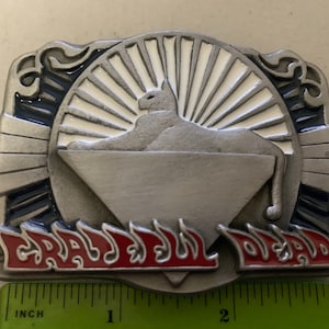 May include: A silver metal belt buckle with a white and blue design. The buckle features a stylized cat lying on a triangle with flames and the text "GRATEFUL DEAD" in red.