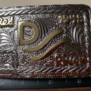 Puede incluir: Una hebilla de cinturón de plata con una 'D' de color dorado y las palabras 'Red River' y '1948' en relieve.