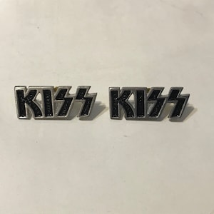 May include: Two metal pins with the word "KISS" in a stylized font. The letters are black with a silver border. The pins are likely for a band or music-related merchandise.