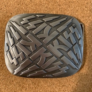 May include: A silver metal belt buckle with a repeating pattern of the word "KISS" in a lightning bolt design.