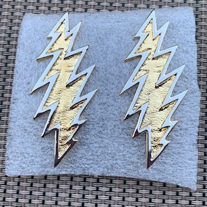 May include: Two gold lightning bolt earrings with a silver outline. The earrings are on a white background.