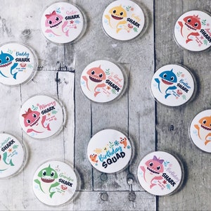 May include: A set of 12 round, white, Baby Shark themed buttons with various family member titles, including "Daddy Shark", "Mommy Shark", "Sister Shark", "Brother Shark", "Cousin Shark", "Auntie Shark", "Uncle Shark", "Grandma Shark", "Grandpa Shark", and "Baby Shark".
