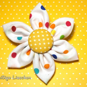 Könnte beinhalten: Eine weiße Stoffblume mit gelber Mitte und bunten Punkten. Die Blume liegt auf einem gelben Hintergrund mit weißen Punkten.