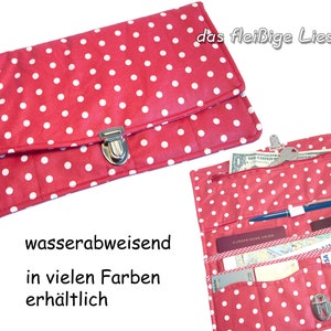 May include: Red and white polka dot wallet with a silver clasp. The wallet is open, revealing multiple compartments for cards, cash, and a pen. The text "wasserabweisend in vielen Farben erhältlich" is visible.