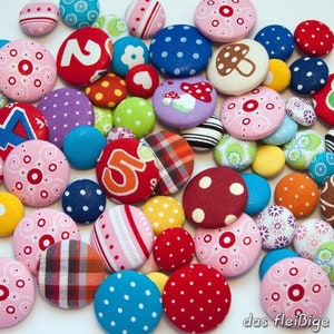 May include: A collection of colorful fabric covered buttons with various patterns and designs, including polka dots, stripes, and floral prints. Some buttons feature numbers, while others have whimsical designs like mushrooms and swirls. The buttons are arranged in a random pattern on a white background.