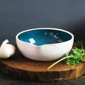 May include: White ceramic bowls with teal and purple interiors are stacked and arranged on a wooden surface. A bulb of garlic and fresh cilantro are also present. The bowls have a modern, minimalist design.