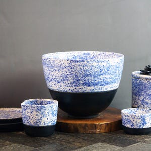May include: A collection of ceramic pottery with a speckled blue and white design, contrasted by a black base. The set includes a large bowl, a small cup, a cylindrical container, and a plate. A dark succulent decorates the container's lid.