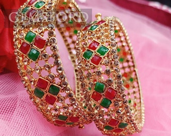 May include: Two gold bangles with a decorative design of clear, red, and green stones. The bangles are on a pink fabric background.