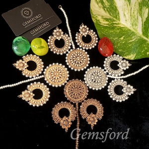 May include: Set of six gold, silver, and bronze Indian bridal jewelry headpieces with pearl accents. Each piece includes a large round pendant with a smaller round earring. The pieces are arranged on a black background with green and red stones.