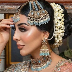 May include: A woman wearing a traditional Indian bridal outfit with a blue and gold headpiece, a matching necklace, and earrings. The headpiece is adorned with pearls and blue stones. The necklace is also adorned with pearls and blue stones. The earrings are long and dangle, with blue stones and pearls.
