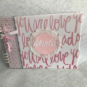Puede incluir: Un álbum de fotos con espiral con una cubierta blanca con texto rosa escrito a mano que dice "you are love". Un círculo rosa con el nombre "Marta" está centrado en la cubierta, rodeado por un tapete de encaje blanco. El lomo tiene una tira decorativa estampada.