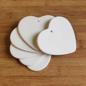May include: A group of white heart-shaped decorations, each with a small hole for hanging. They are arranged in a fan shape on a wooden surface. The hearts have a smooth, matte finish, perfect for crafting or decoration.