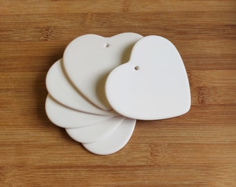 May include: A collection of white heart-shaped ornaments with a small hole at the top for hanging. The ornaments are arranged in a fan shape, resting on a wooden surface. The hearts have a smooth, matte finish.