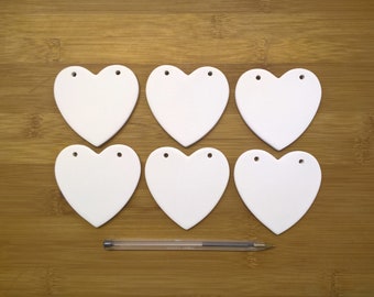 May include: Six white ceramic heart-shaped ornaments with holes for hanging. The ornaments are arranged in two rows of three on a wooden surface.