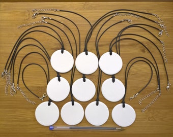 May include: Ten blank white round ceramic pendants with black cords and silver chains. The pendants are arranged in a circle on a wooden surface.