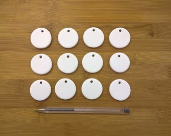 May include: Twelve white ceramic round ornaments with a hole in the center. The ornaments are arranged in three rows of four on a wooden surface.