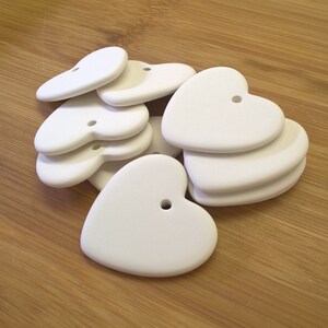May include: A pile of white ceramic heart-shaped ornaments with a hole in the centre of each heart. The hearts are arranged in a random pattern on a wooden surface.