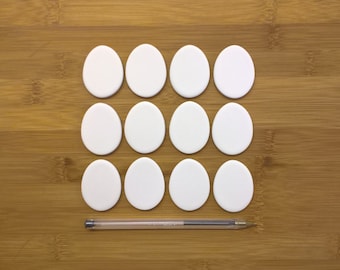 May include: Twelve white, oval-shaped objects arranged in a grid pattern on a wooden surface. A clear pen with a black ink cartridge lies horizontally below the objects. The objects appear to be made of a smooth, solid material.