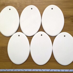 May include: Six white ceramic oval ornaments with a hole for hanging. The ornaments are blank and ready to be painted or decorated.