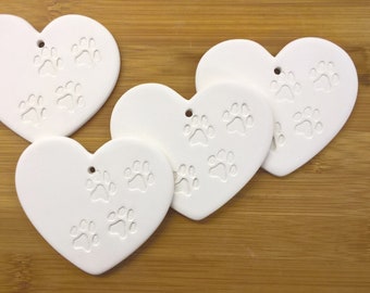 May include: Four white ceramic heart-shaped ornaments with paw print designs. The ornaments have a hole at the top for hanging.