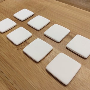 May include: Eight white square ceramic coasters on a wooden surface. The coasters are smooth and have a matte finish.