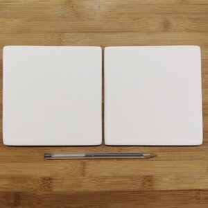 May include: Two blank white square ceramic tiles on a wooden surface. A silver pen is lying between the tiles.