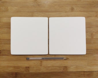 May include: Two blank white square ceramic tiles on a wooden surface. A silver pen is lying between the tiles.