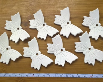 May include: Eight white ceramic butterfly-shaped ornaments with a hole in the center. The ornaments are arranged on a wooden surface.