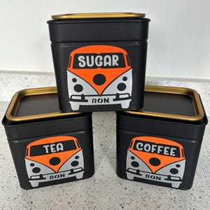 May include: Three black metal storage tins with gold-coloured lids. Each tin features a retro orange and white graphic of a van with the words "SUGAR", "TEA", and "COFFEE". The word "RON" is printed below the van graphic.