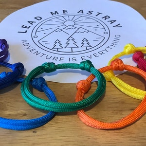 May include: A set of six colourful friendship bracelets in a rainbow pattern. The bracelets are made of woven cord and have a knot at the end. The bracelets are arranged in a circle on a wooden surface. The text "Lead Me Astray Adventure Is Everything" is visible on a white circle in the centre of the image.