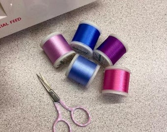 Könnte beinhalten: Fünf Garnrollen in verschiedenen Farben, darunter rosa, lila, blau und ein hellerer Blauton. Ein Paar pinkfarbener Scheren ist ebenfalls auf dem Bild zu sehen.