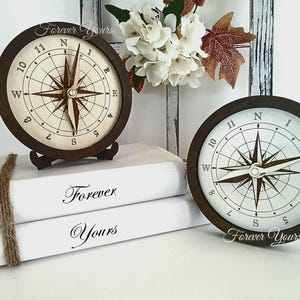 May include: Two wooden compass clocks with a white background and brown trim. The compass needles point north. The clocks are on a stack of white books with the words "Forever Yours" written on them.