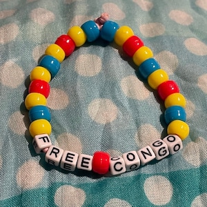 Könnte beinhalten: Ein Perlenarmband mit der Aufschrift "FREE CONGO". Das Armband hat abwechselnde Reihen aus gelben, roten und blauen Perlen. Die Buchstaben befinden sich auf weißen quadratischen Perlen, mit einer roten Perle, die die Wörter trennt.