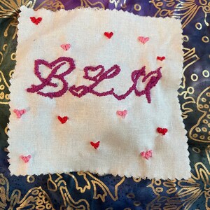 May include: A white fabric square embroidered with the word "BLU" in dark pink cursive. Small red and pink hearts are scattered around the letters. The background is a patterned fabric with gold and blue tones.