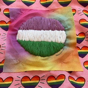 May include: A square of fabric with a gradient of colors, featuring an embroidered design of lips in purple, white, and green. The background is pink fabric with rainbow heart patterns, creating a vibrant and colorful composition.