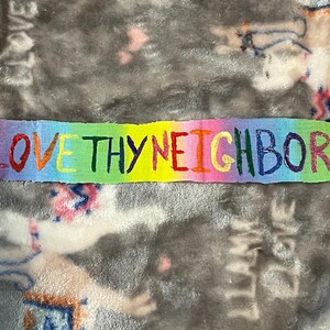 May include: A rectangular fabric patch with the words "LOVE THY NEIGHBOR" in rainbow-colored letters. A small red heart is next to the word "NEIGHBOR". The patch is on a gray and white patterned surface.