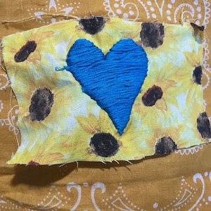 Puede incluir: Un parche con un corazón azul bordado sobre tela amarilla con estampado de girasoles. El parche está cosido sobre un pañuelo bandana amarillo mostaza con un diseño de cachemir blanco. El corazón es el punto focal, con los girasoles de fondo.