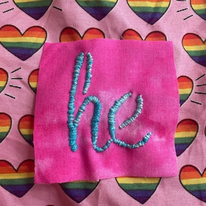 May include: A pink fabric square embroidered with the word "he" in turquoise thread. The background is a pink fabric patterned with rainbow-colored hearts.