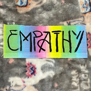 Puede incluir: Un parche rectangular con la palabra "EMPATHY" en letras negras, estilo manuscrito. El fondo es un degradado de colores arcoíris, incluyendo rosa, azul, verde y amarillo. El parche está sobre una superficie gris y estampada.