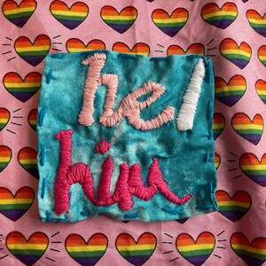 May include: A turquoise fabric patch embroidered with the words "he/him" in pink and white thread. The patch is stitched with blue thread and set against a pink background patterned with rainbow hearts.