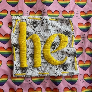 May include: Pink fabric with rainbow hearts and a square patch. The patch has the word "be" embroidered in yellow thread. The patch has a white and black pattern with bees and jars.