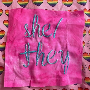 Może przedstawiać: Różowy kwadrat materiału z wyhaftowanymi słowami "she/they" turkusową nicią. Tło to różowy materiał we wzór tęczowych serc.