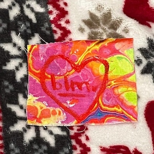 May include: A small, rectangular fabric patch with a vibrant, abstract design in shades of pink, orange, yellow, and blue. A red heart shape with the letters "blm" is drawn on top. The patch is placed on a patterned background.
