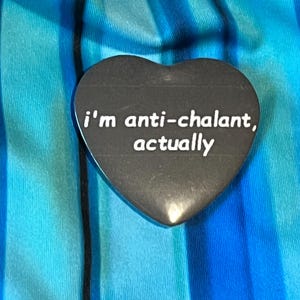 May include: A black heart-shaped pin with the phrase "i'm anti-chalant, actually" in white lettering. The pin is set against a background of blue and teal fabric with vertical stripes.