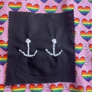 May include: A black fabric square featuring two white embroidered anchor designs. The square is set against a pink background patterned with rainbow-coloured hearts. The edges of the black square are unfinished.