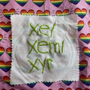 Op de afbeelding: Een witte stoffen vierkant met groene geborduurde tekst "xel xeml xyr" tegen een roze achtergrond met regenboogkleurige harten. De harten hebben stralende lijnen en de stof heeft een rafelige rand.