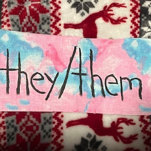 Könnte beinhalten: Ein rechteckiges Schild mit den Worten "they/them" in schwarzer Tinte geschrieben. Der Hintergrund ist eine Mischung aus rosa und blauer Farbe im Aquarellstil. Das Schild befindet sich auf einer gemusterten Decke mit roten und weißen Rentieren.