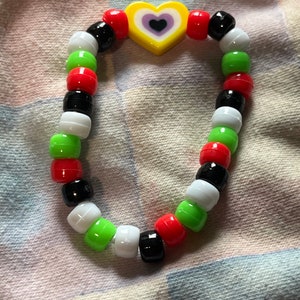 May include: A beaded bracelet featuring a yellow heart-shaped bead with a purple heart in the center. The bracelet is made of red, black, white, and green beads. The bracelet is on a patterned fabric background.
